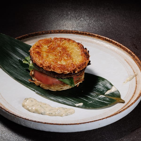 Kogane Ramen's ramen burger with crispy noodle buns, patty, tomato, and lettuce on a leaf.